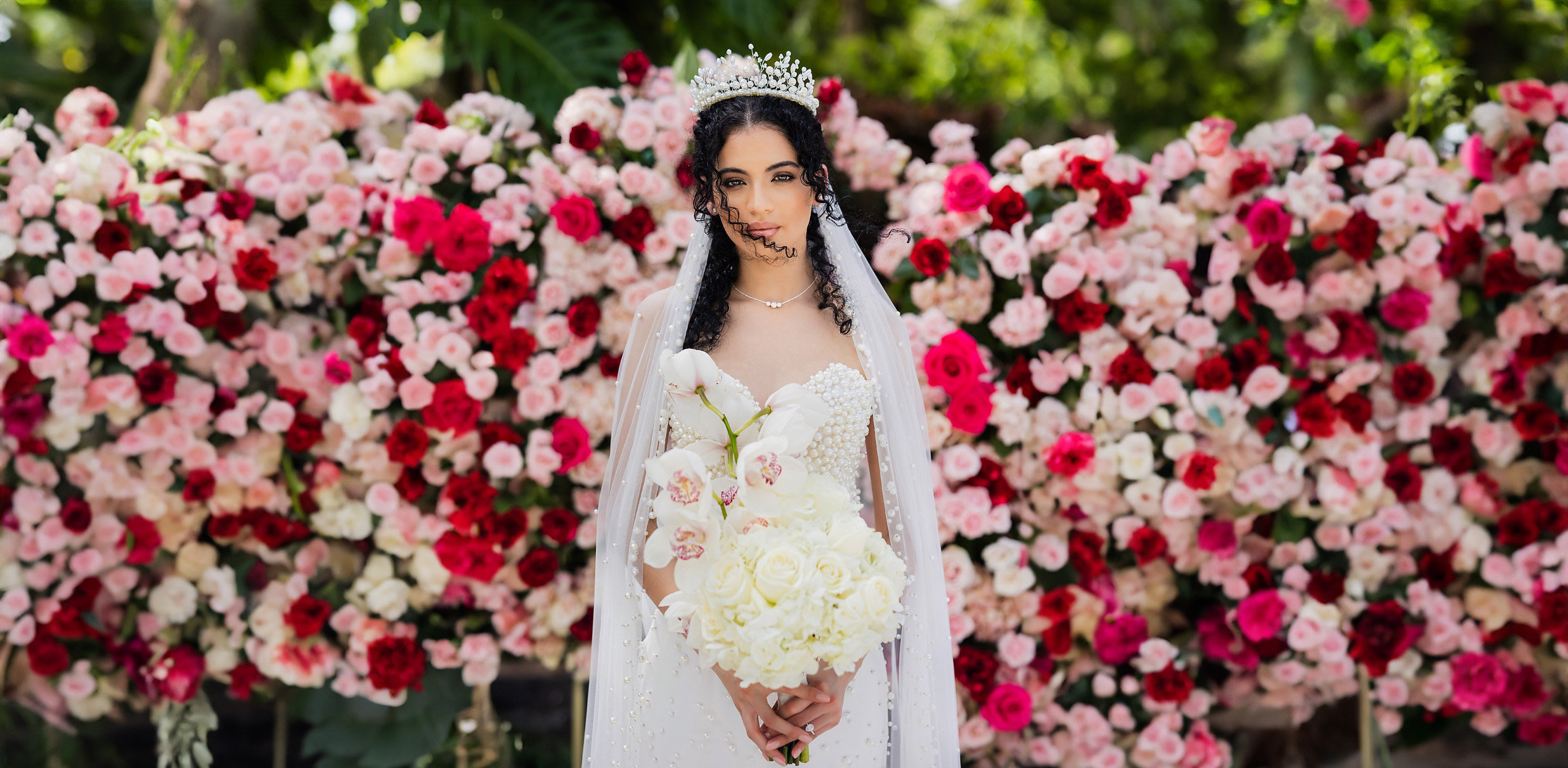Bride in a pearl crown holding a white orchid bouquet before a wall of pink and red roses