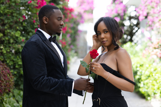 Elegant couple in black attire with a red rose among bougainvillea